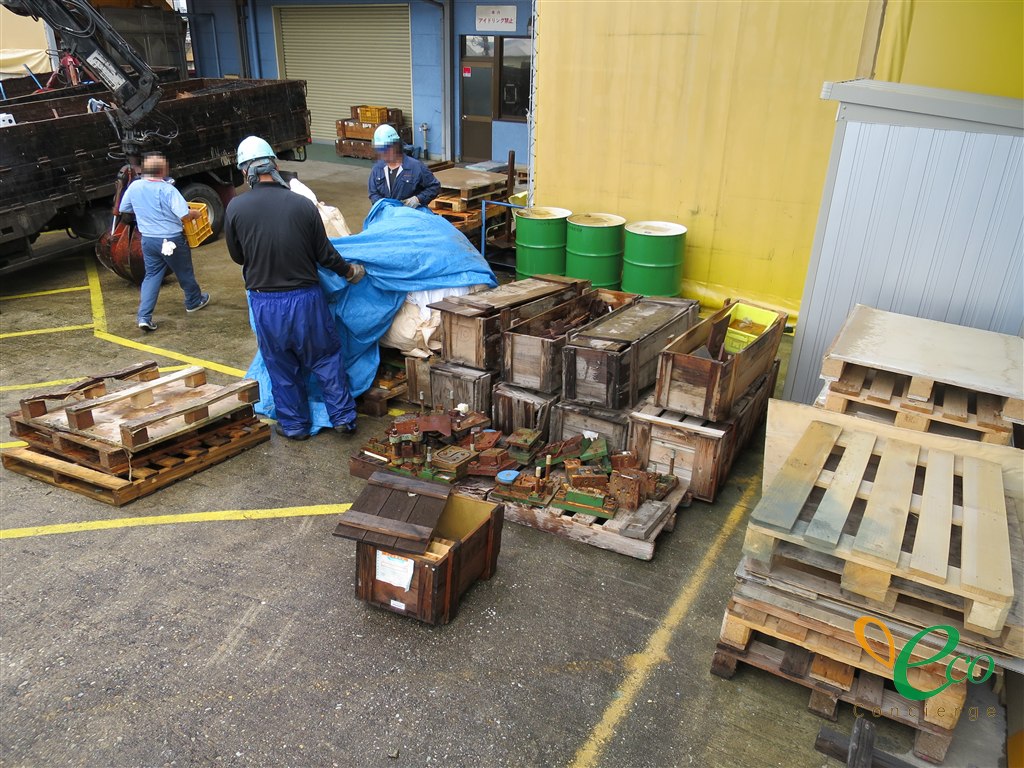 屋外のヤードで、クレーン付きトラックの手前に木箱と木製パレットが並び、パレットの上に使用済みの金型が置かれている様子