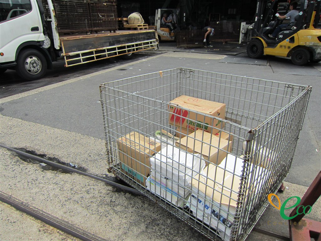 屋外の作業場に、書類を積んだ金網カゴが置かれ、奥ではトラックやフォークリフトによる荷下ろし作業が行われている様子