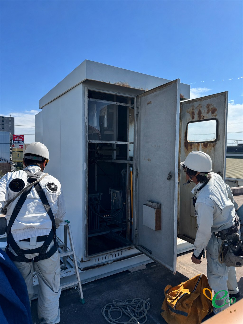 屋上に設置されたキュービクルの扉を開放し、フルハーネスを装着した作業員2名が作業している様子
