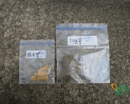 灰色のざらついた床の上に並べられた2つの透明な袋。それぞれに「給水管」「排気管」と手書きされている