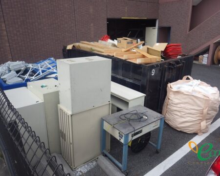 黒い金属製の箱に木材や段ボールが積まれ、手前に事務用のキャビネットや机が並んでいる