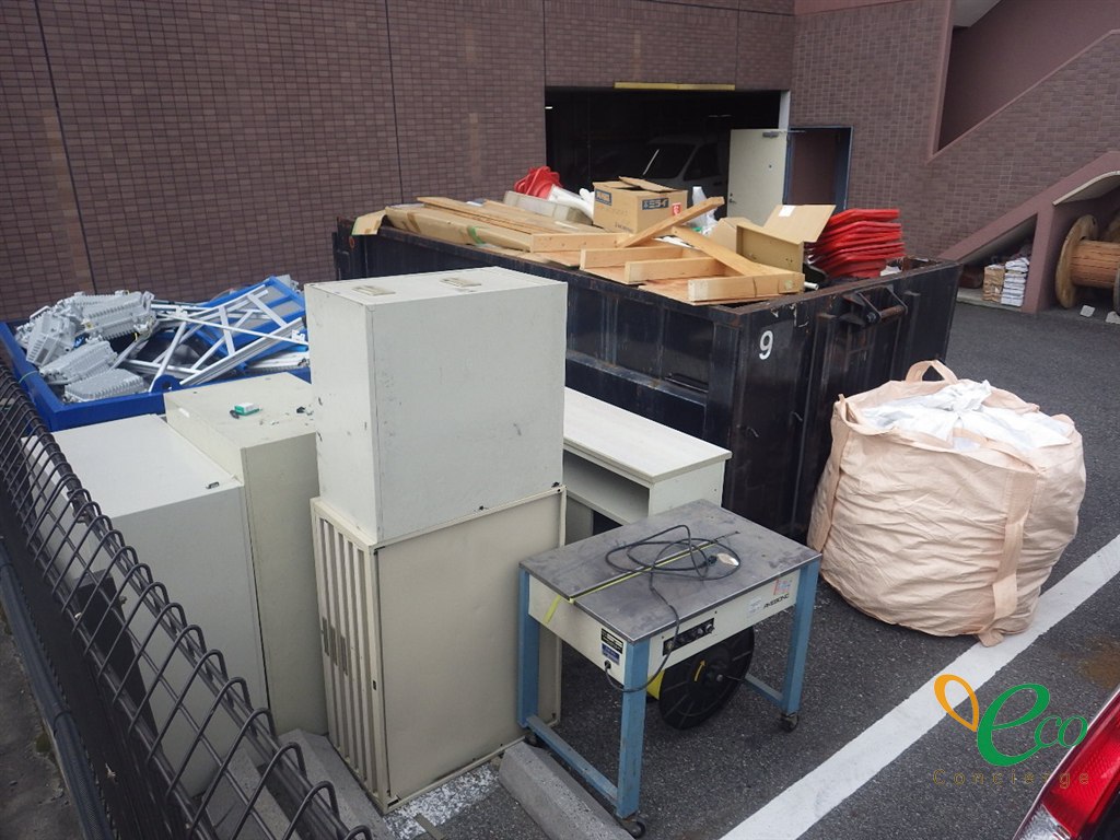 黒い金属製の箱に木材や段ボールが積まれ、手前に事務用のキャビネットや机が並んでいる