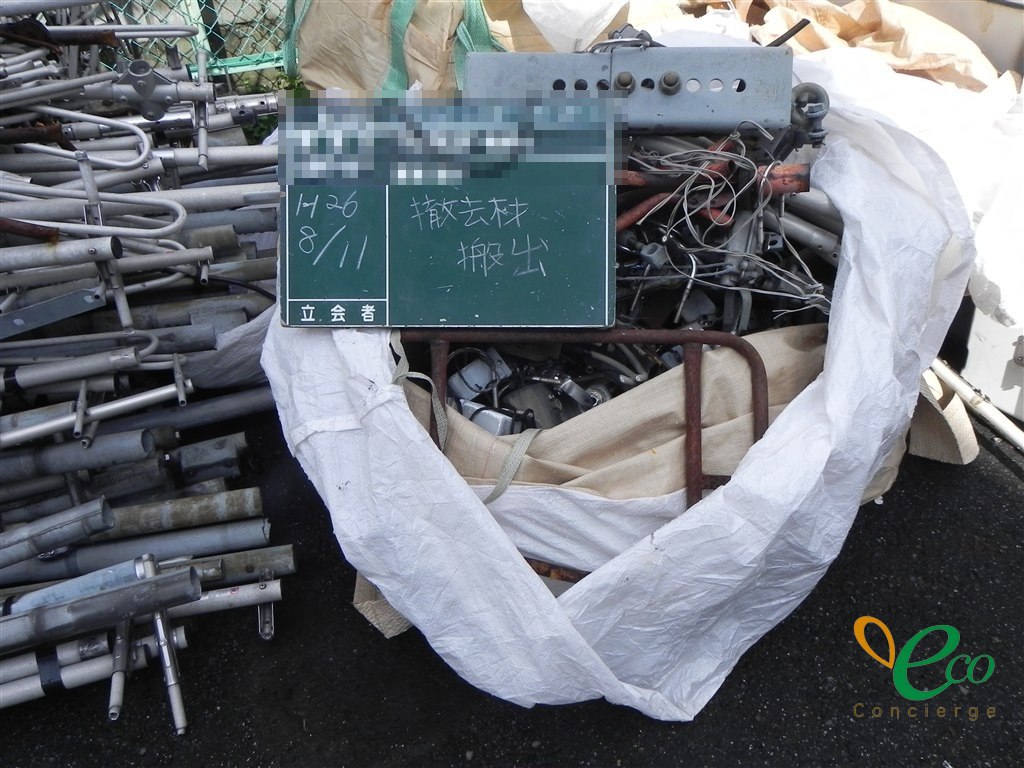 めっきが施された鋼管と取付金具が積み上げられ、隣の白いフレコンバッグに機器の筐体とみられる灰色の箱と赤く錆びた管が入っている様子。工事記録用の黒板の記載内容にはモザイク処理を施している