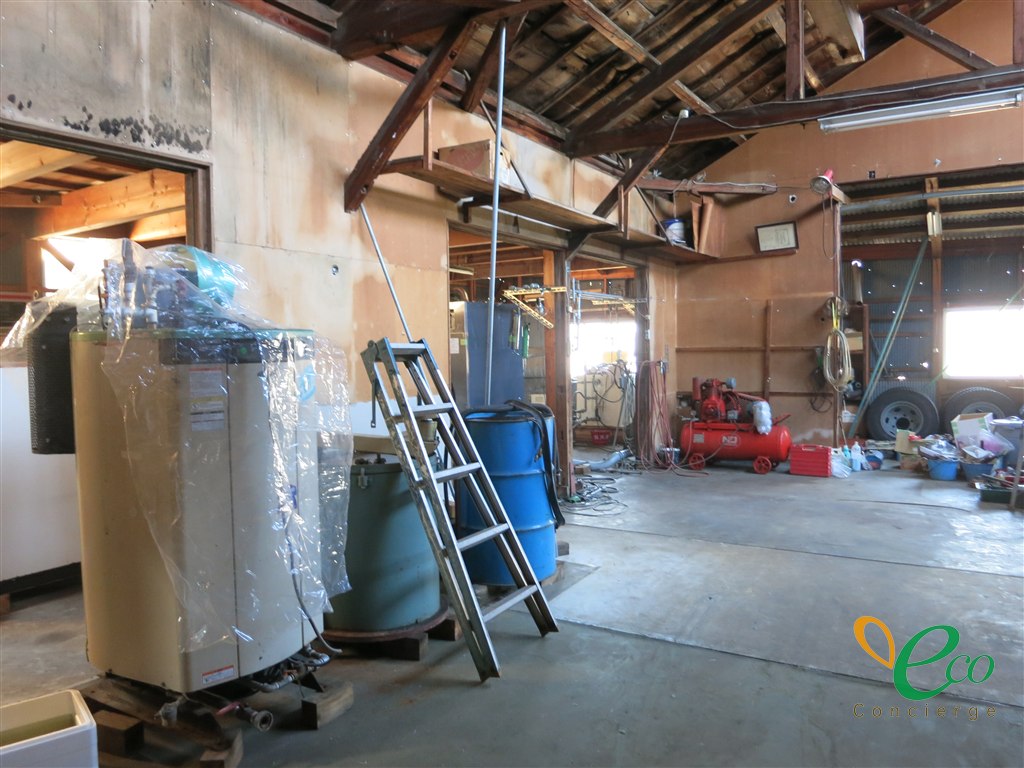建屋の中に、ビニールで養生された縦型の機器、緑と青のドラム缶、脚立が置かれ、奥の壁ぎわに赤いエアコンプレッサーが見える様子