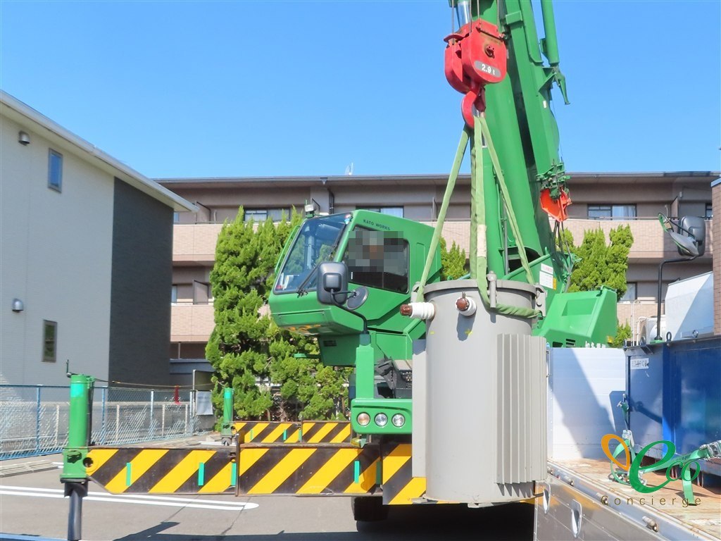 緑色のクレーン車の正面。円筒形の変圧器が緑色のスリングで吊られ、右手前のトラック荷台の近くまで運ばれてきた様子。運転席に作業員が座っている