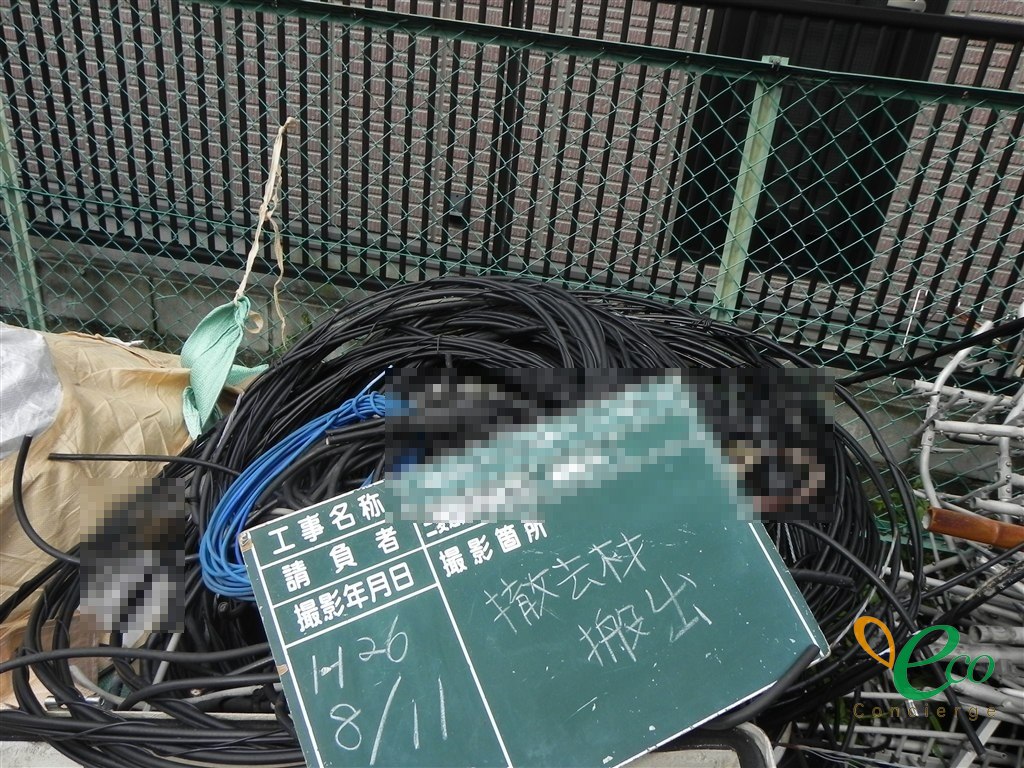 黒と青の通信用とみられるケーブルがコイル状に巻かれ、資材置き場の地面に置かれている。奥に緑色のフェンスと建物が見える。工事記録用の黒板の記載内容にはモザイク処理を施している