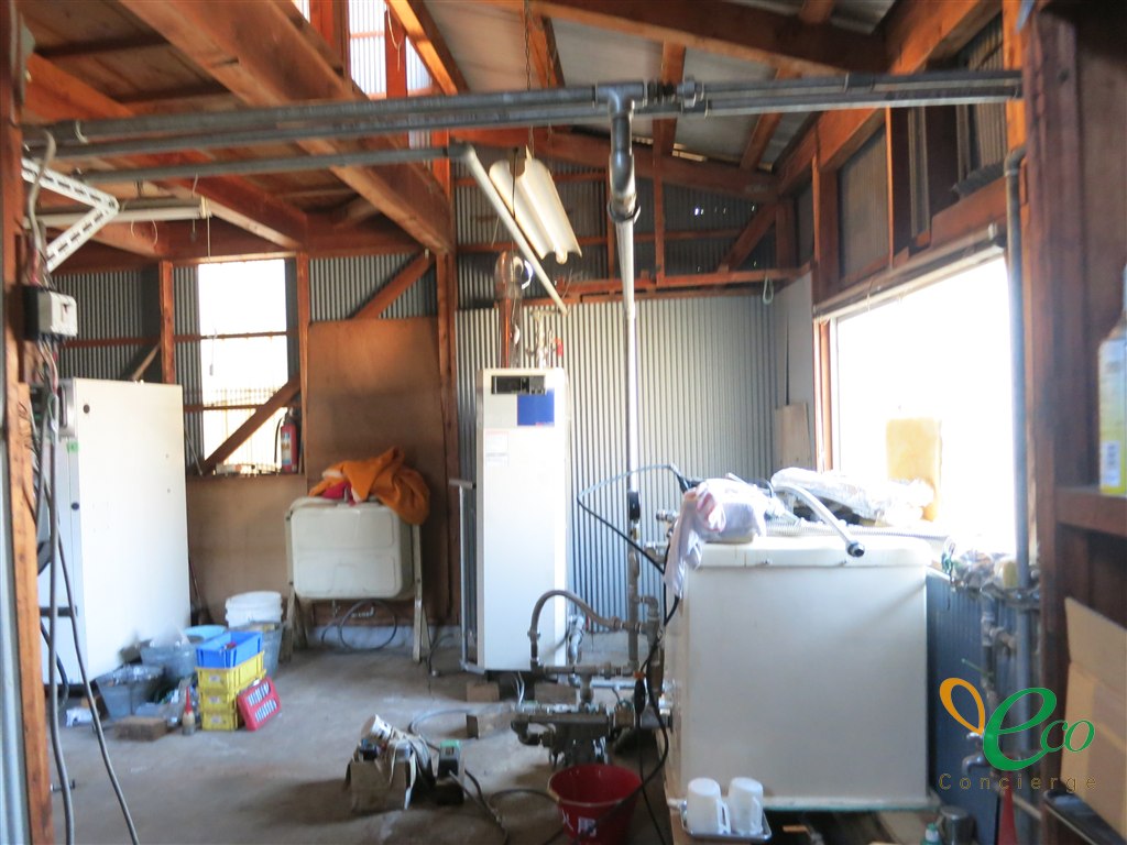 木造の建屋の中に、白い縦型の機器と白い箱型の設備が置かれ、天井から床へ鋼管の配管とバルブが伸びている様子