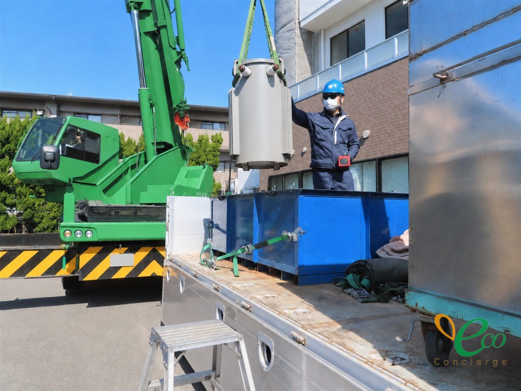 トラック荷台の上で、青い収納箱の近くまで吊り下ろされた円筒形の変圧器に、作業員が手を添えている様子