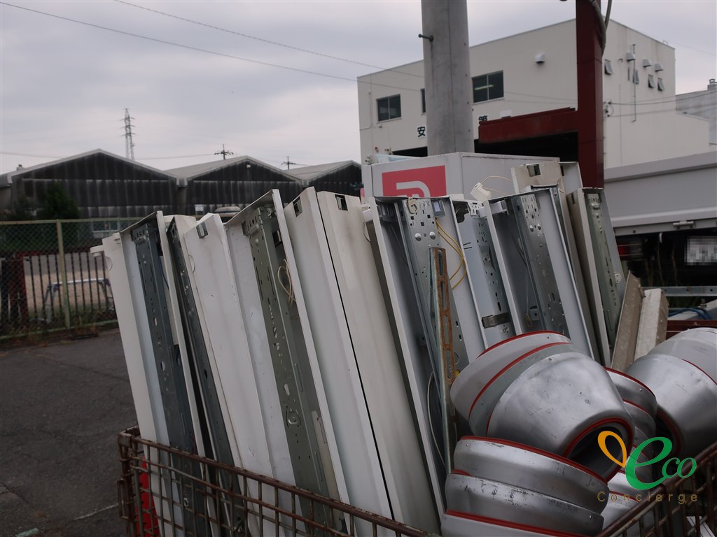 金属製のカゴに、照明器具の筐体・反射板が立てた状態で並べられ、手前に円筒状の金属部材が置かれている様子