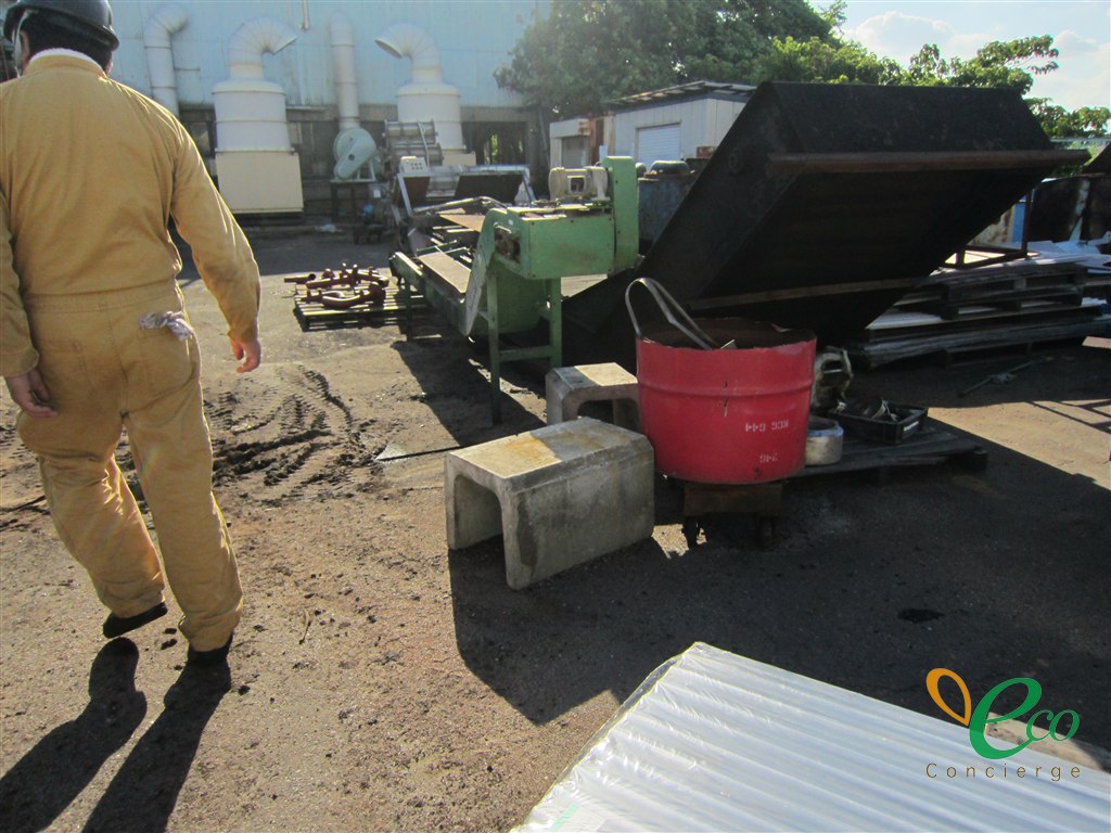 作業服とヘルメットを着けた人が、後ろ姿で工場の敷地内を歩いている様子。奥に白い建物と配管、緑色の機械、鉄製の箱、赤いドラム缶が見える
