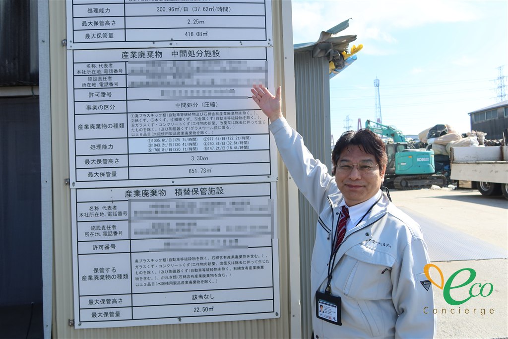 株式会社エコ・コンシェルジュの作業着を着た代表取締役が、産業廃棄物中間処分施設の掲示板を指さしている様子。掲示板には事業の区分、産業廃棄物の種類、処理能力、最大保管量が並んでいる