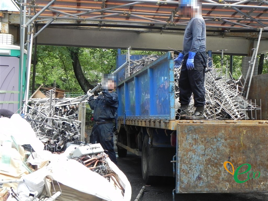 作業員2名が、鋼管をトラックの荷台に積み込んでいる様子。1名が荷台の上に立ち、もう1名が下から鋼管を差し上げている。顔とヘルメットの表示にはモザイク処理を施している