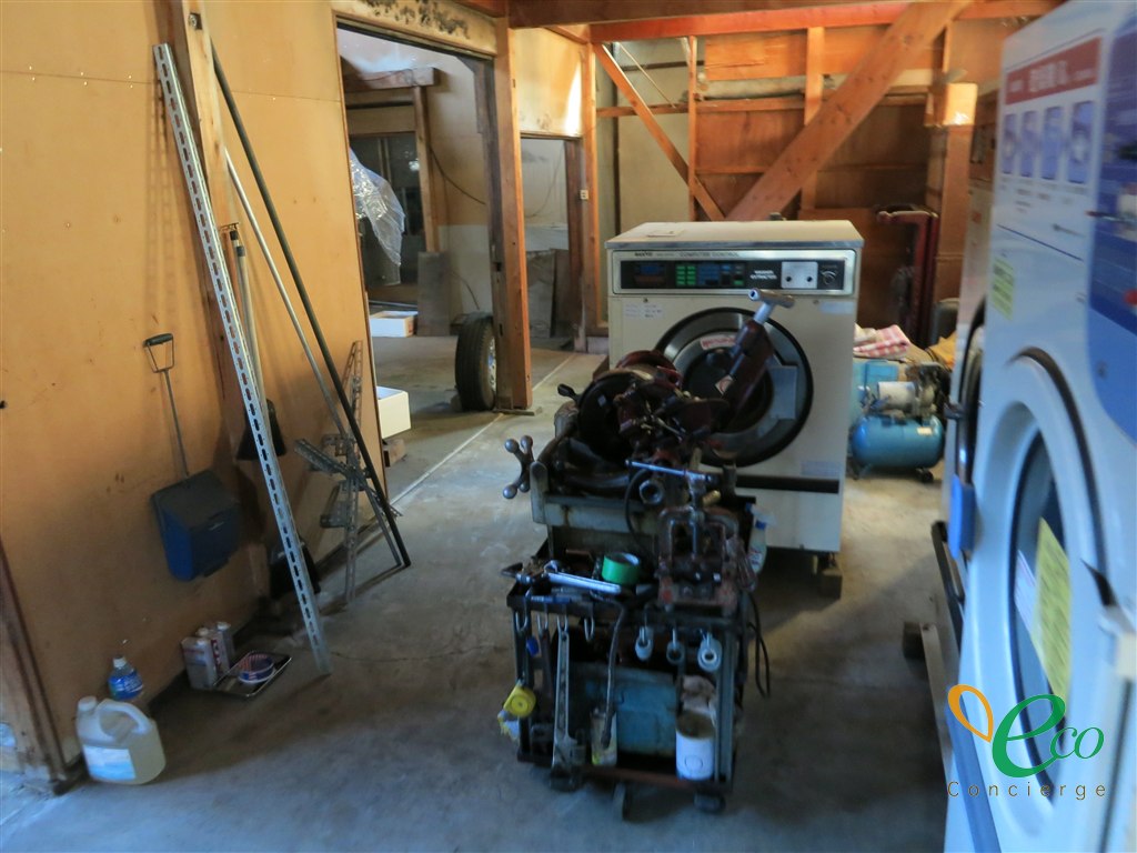 建屋の中に業務用洗濯機が置かれ、その手前に配管加工用とみられる工具を載せた台車、右奥に水色のタンク式コンプレッサーがある様子