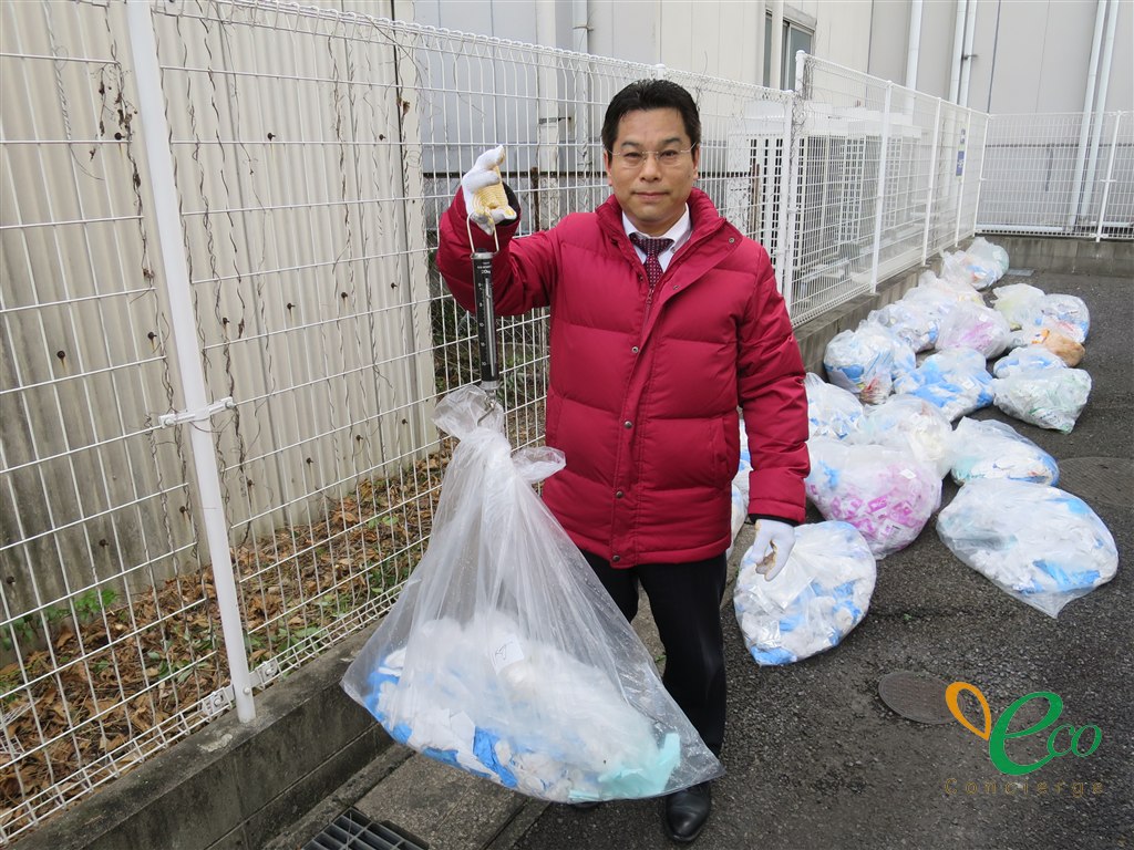 作業用手袋をした担当者が、吊り下げ式の秤で透明なポリ袋を持ち上げて重さを確認している様子。奥の通路には同じような袋がいくつも並んでいる