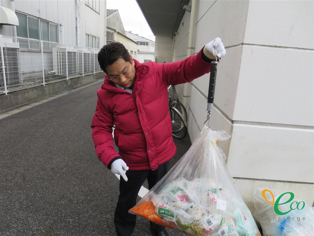 担当者が吊り下げ式の秤で透明なポリ袋を吊り上げ、袋の中を見ている様子。袋の中には食品の残りと使用済みの容器・包装材が混ざって入っている