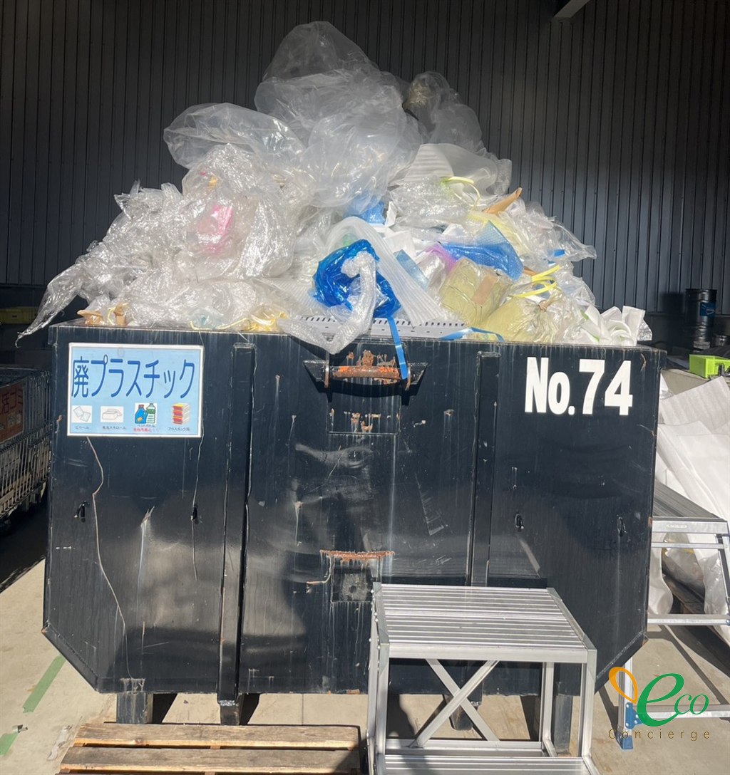 「廃プラスチック」の表示と「No.74」の番号がある黒いコンテナから、ビニールや袋があふれている様子