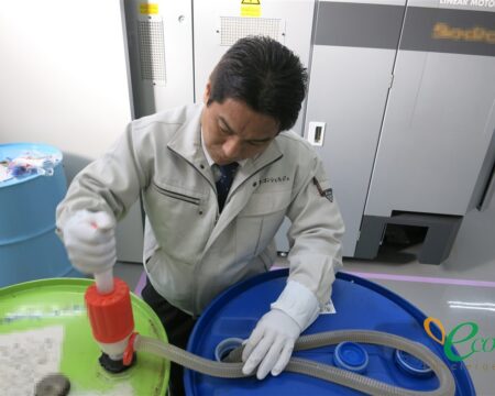 作業着姿の人物が、緑色のドラム缶からポンプで青いドラム缶へ油を移し替えている様子