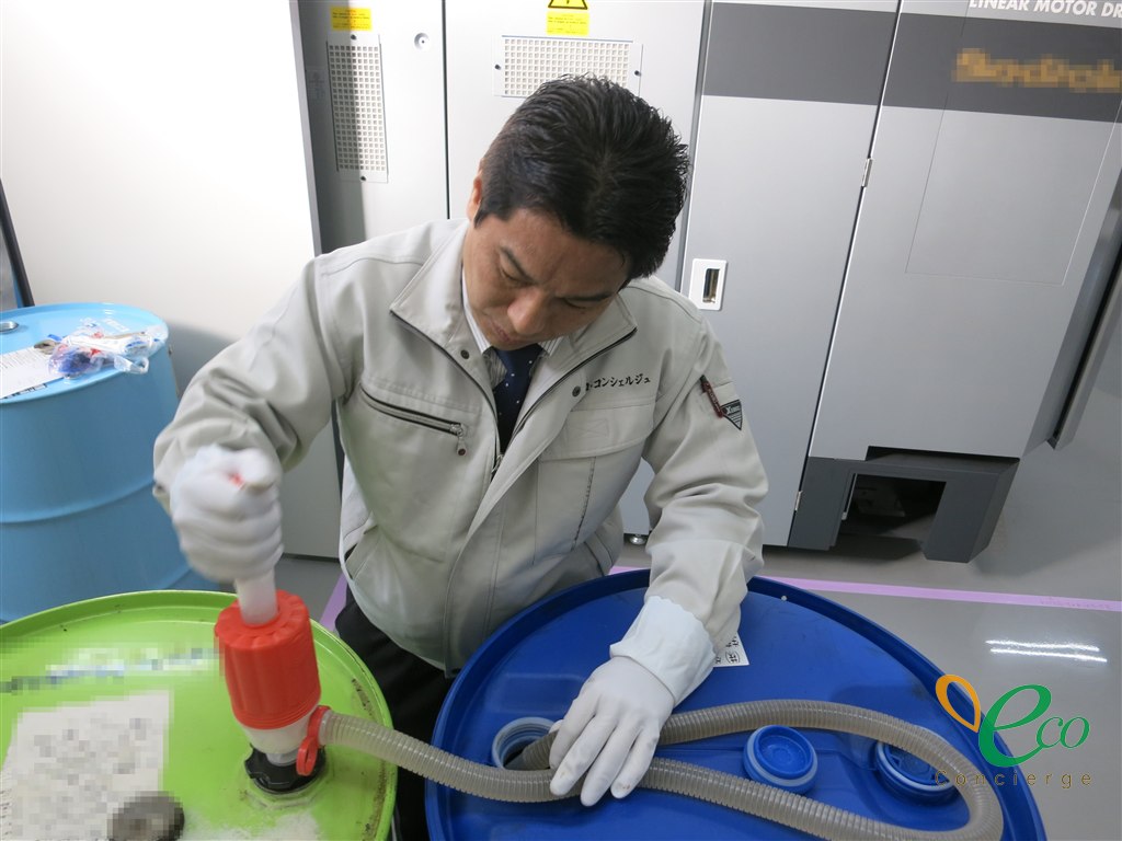 作業着姿の人物が、緑色のドラム缶からポンプで青いドラム缶へ油を移し替えている様子