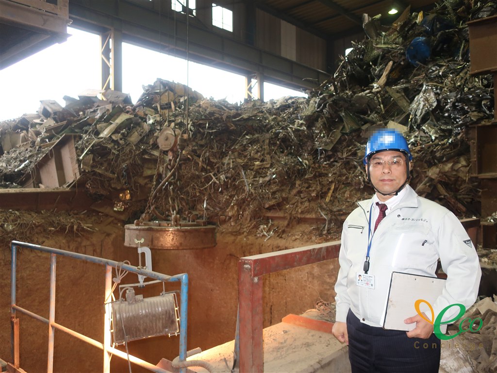 作業着姿の代表取締役が、鉄スクラップの山を背に立っている様子。ヘルメット正面のロゴはモザイク処理済み