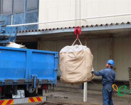 青いトラックからクレーンで大きな袋を吊り上げ、作業員が荷下ろしの作業をしている様子。作業員の顔・ヘルメットの社名・トラックのナンバーはモザイク処理済み