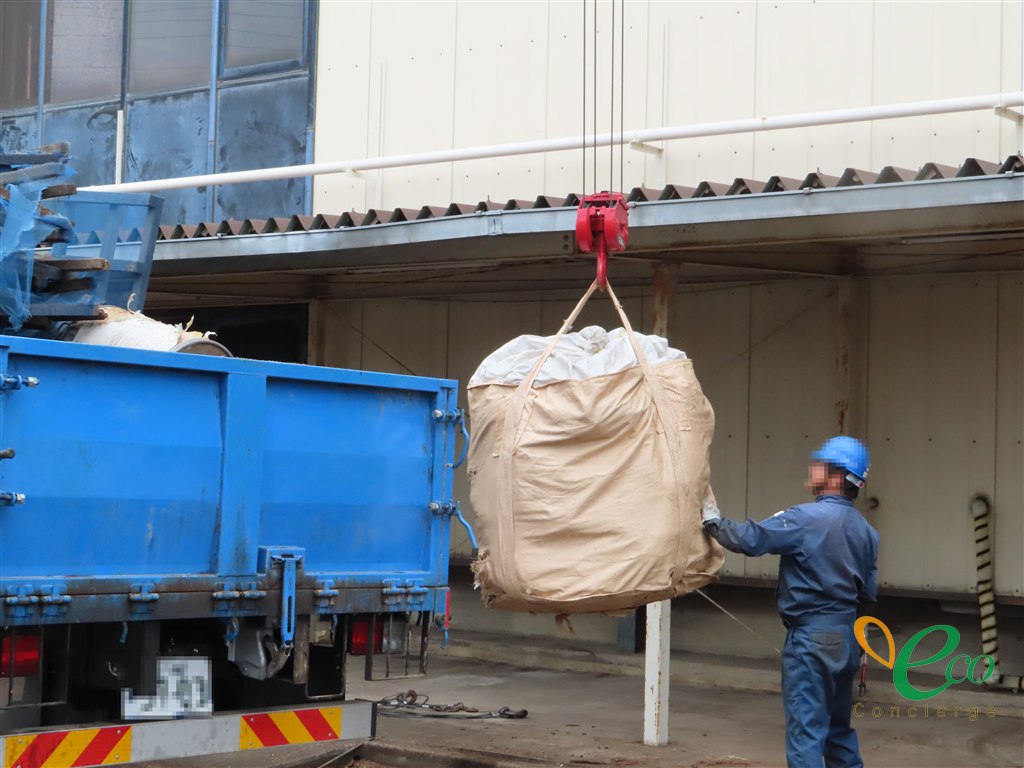 青いトラックからクレーンで大きな袋を吊り上げ、作業員が荷下ろしの作業をしている様子。作業員の顔・ヘルメットの社名・トラックのナンバーはモザイク処理済み