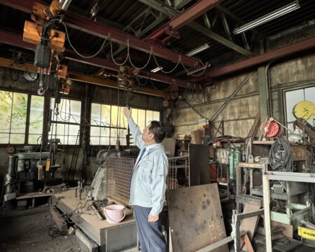 作業着姿の人物が、工場内でクレーン設備を見上げて指さしている様子。古い機械や資材が周囲に置かれている
