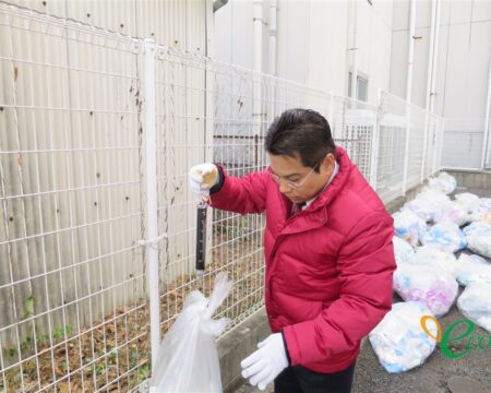 担当者が吊り下げ式の秤を持ち、袋の重さを確認している様子。右手の地面には袋が並んでいる
